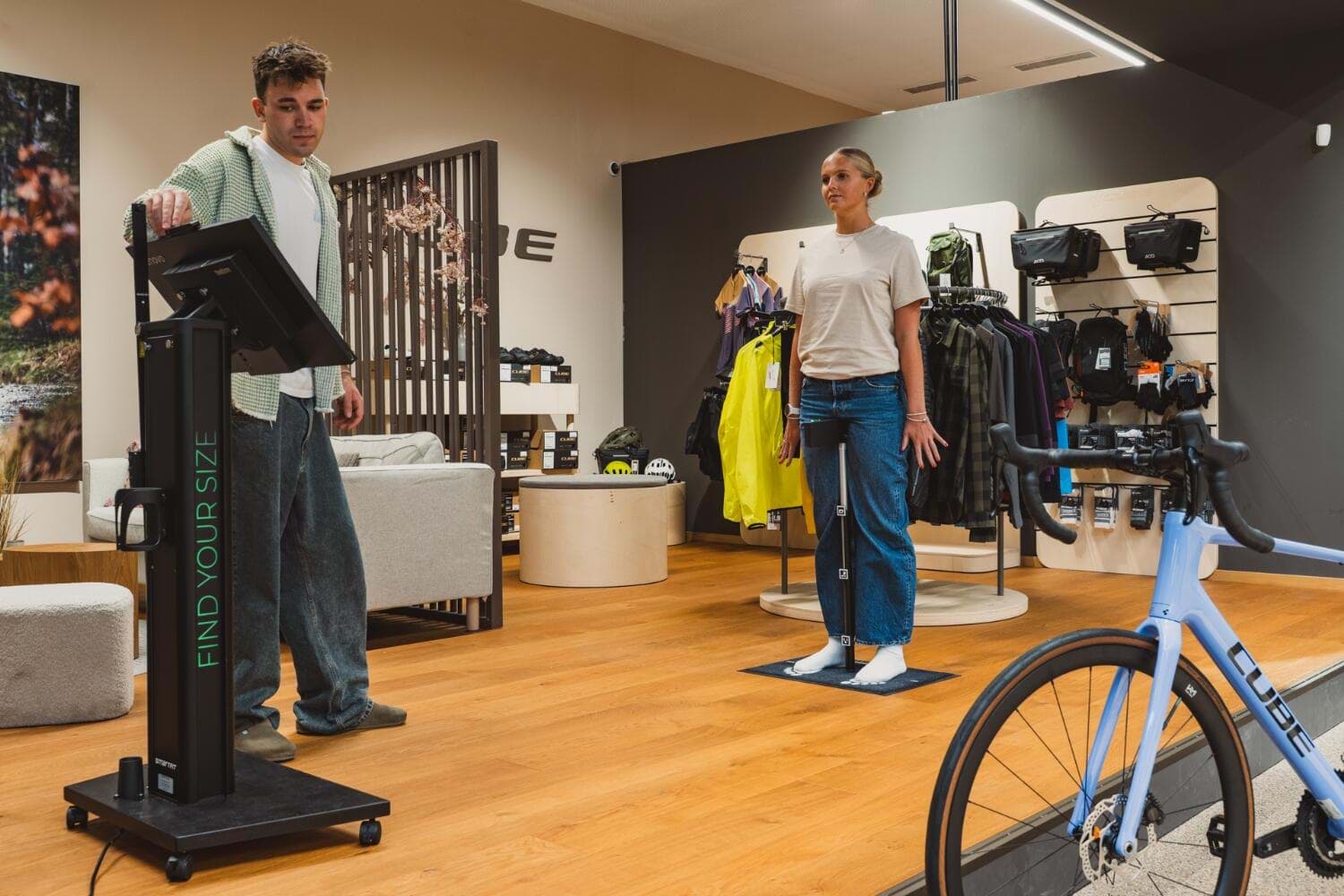 Beratung an der SmartFit-Messsäule im Store, daneben eine Kundin auf der Messmatte und ein Rennrad