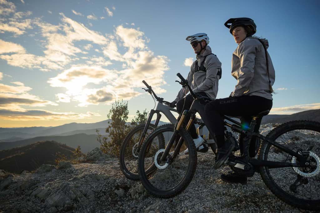 Zwei Radfahrer auf einem Felsgrat blicken in der Abendsonne ins Tal